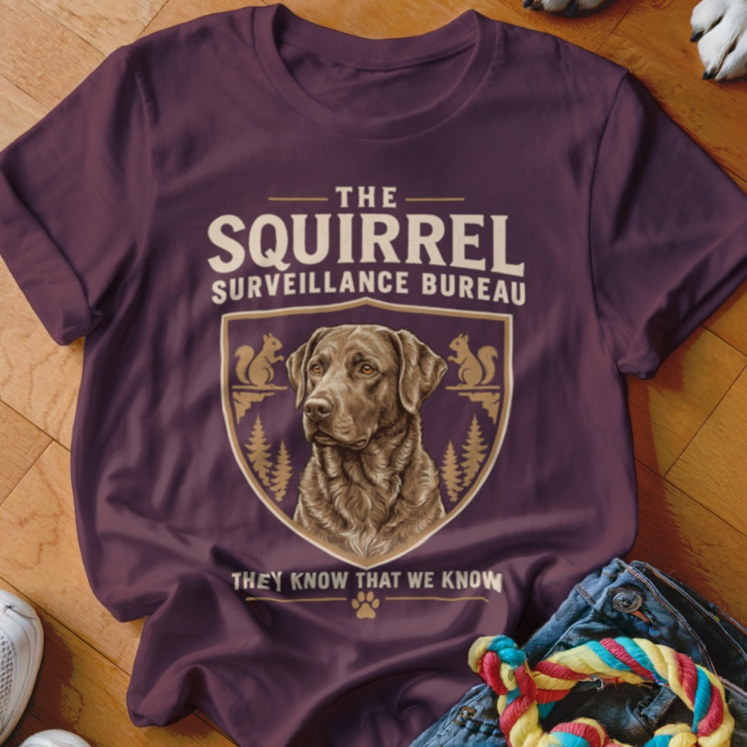 Plum-colored t-shirt featuring a graphic of a chocolate Labrador and text reading The Squirrel Surveillance Bureau.