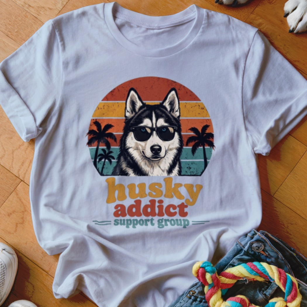 A white t-shirt featuring a retro graphic of a husky wearing sunglasses with sunset and palm trees, and text reading husky addict support group.