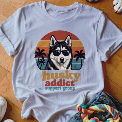 A white t-shirt featuring a retro graphic of a husky wearing sunglasses with sunset and palm trees, and text reading husky addict support group.