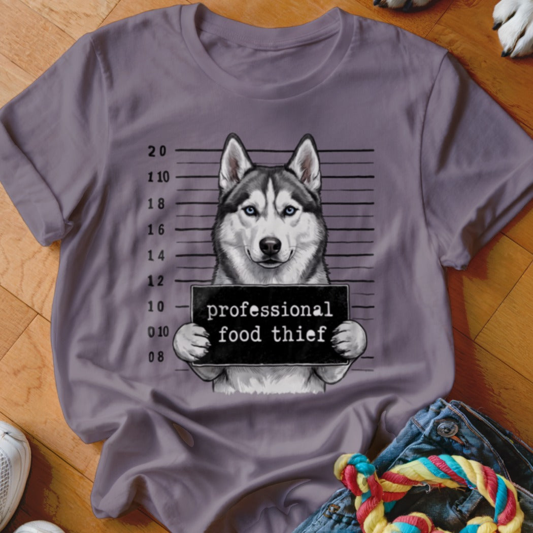 Husky dog mugshot with "professional food thief" sign on purple t-shirt.