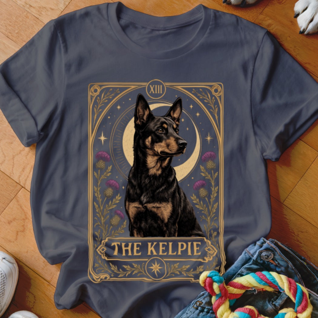 A dark gray t-shirt with a tarot card graphic featuring a Kelpie dog, moon, thistles, and text reading THE KELPIE.